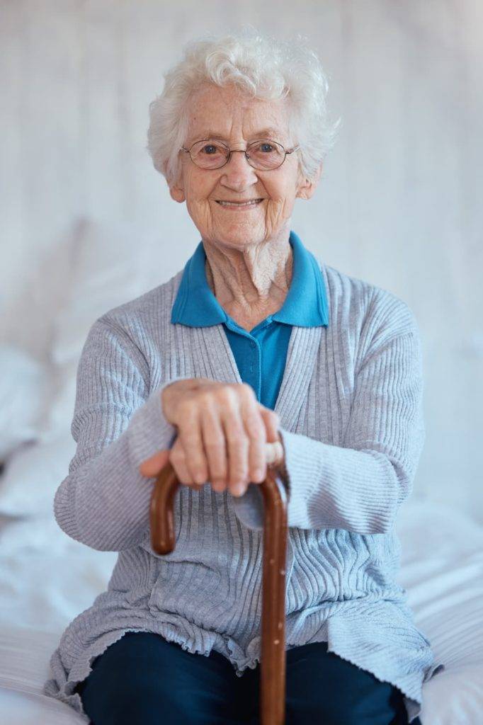 Elderly lady sat on a bed with a walking stick in her hands